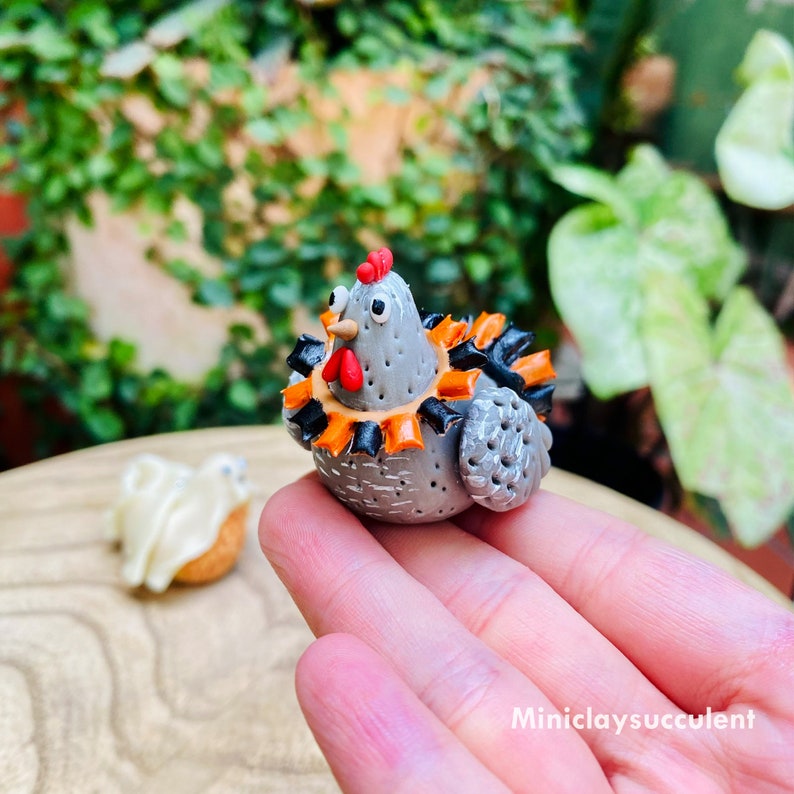 May include: A gray and white ceramic chicken figurine with orange and black accents. The chicken is wearing a festive orange and black collar with a red beak and eyes.