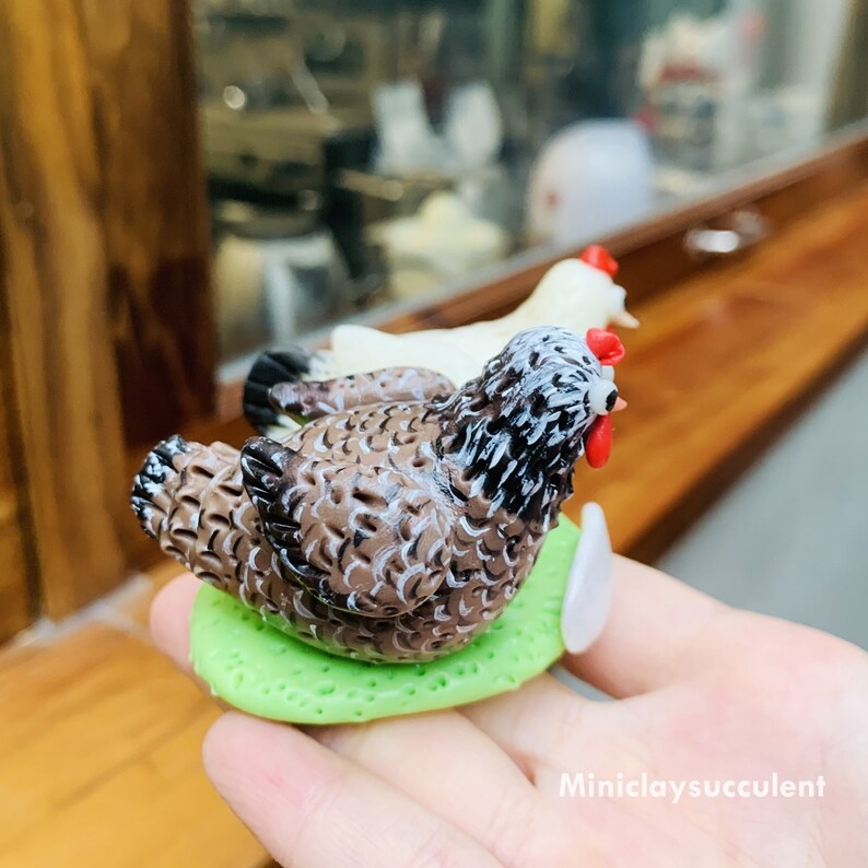 Peut inclure: Une figurine de poulet brun et blanc avec une cr&ecirc;te rouge assise sur une feuille verte. Le poulet est perch&eacute; sur une base blanche.