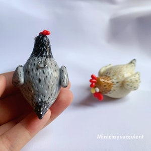 Peut inclure: Deux figurines de poulets en p&acirc;te polym&egrave;re faites &agrave; la main. L'une est grise avec des mouchetures noires et une cr&ecirc;te rouge, l'autre est blanche avec des mouchetures brunes et une cr&ecirc;te rouge. Les deux ont une petite boucle argent&eacute;e au dos.