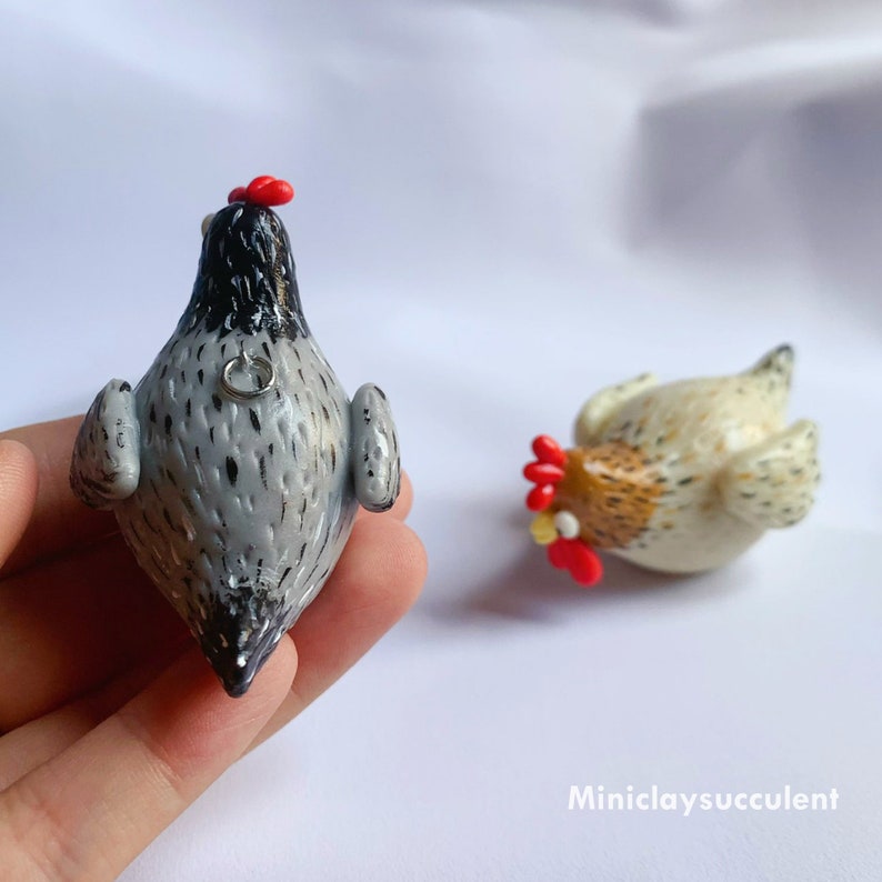 Peut inclure: Deux figurines de poulets en p&acirc;te polym&egrave;re faites &agrave; la main. L'une est grise avec des mouchetures noires et une cr&ecirc;te rouge, l'autre est blanche avec des mouchetures brunes et une cr&ecirc;te rouge. Les deux ont une petite boucle argent&eacute;e au dos.
