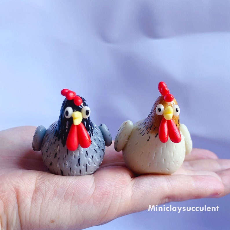 Peut inclure: Deux poulets en argile polym&egrave;re faits &agrave; la main. L'un est gris avec des accents noirs et l'autre est blanc avec des accents bruns. Les deux ont des cr&ecirc;tes et des barbillons rouges.