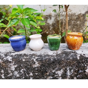 Puede incluir: Cuatro pequeñas macetas de cerámica en diferentes colores: azul, blanco, verde y naranja. Las macetas están dispuestas en fila sobre una superficie de piedra. MiniClaySucculent