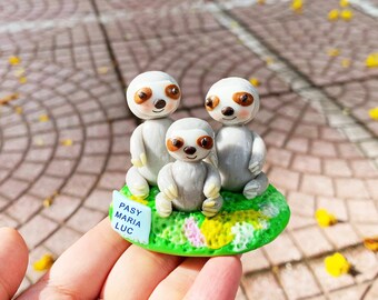 Puede incluir: Tres adorables figuritas de perezosos hechas de arcilla polimérica, sentadas sobre una base verde con flores amarillas y rosas. La base tiene un pequeño cartel azul que dice "Pasy Maria Luc".