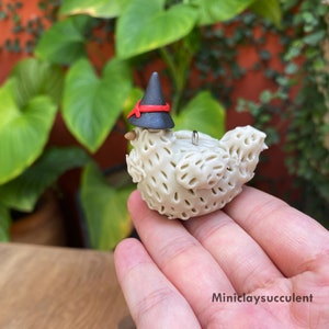 May include: A white ceramic chicken figurine wearing a black witch hat with a red band. The chicken has a textured surface and a small loop on its back.