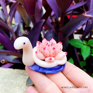 May include: A handmade polymer clay snail sculpture featuring a pink succulent with red tips. The snail has a white body, a blue base, and the text "MINICLAYSUCCULENT". The sculpture is designed to resemble a succulent.