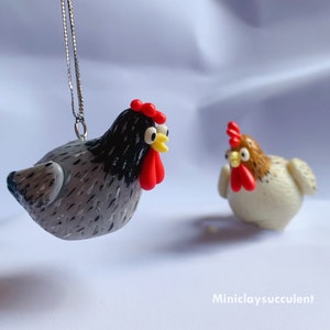 Peut inclure: Deux d&eacute;corations de poulet en p&acirc;te polym&egrave;re faites &agrave; la main. L'une est grise avec des marques noires et une cr&ecirc;te rouge, l'autre est blanche avec des marques brunes et une cr&ecirc;te rouge. Les deux ont des yeux jaunes.