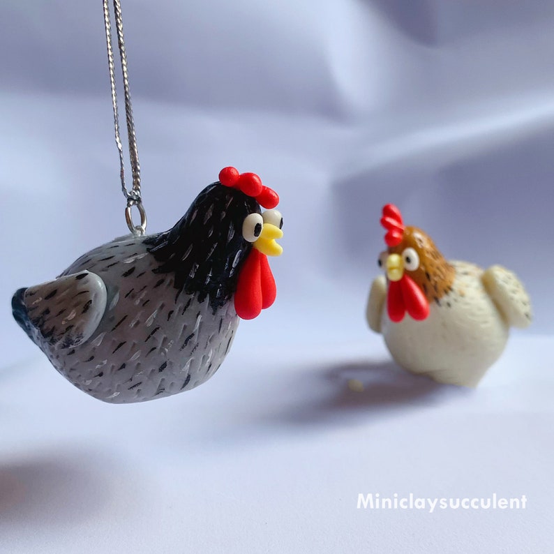 Peut inclure: Deux d&eacute;corations de poulet en p&acirc;te polym&egrave;re faites &agrave; la main. L'une est grise avec des marques noires et une cr&ecirc;te rouge, l'autre est blanche avec des marques brunes et une cr&ecirc;te rouge. Les deux ont des yeux jaunes.