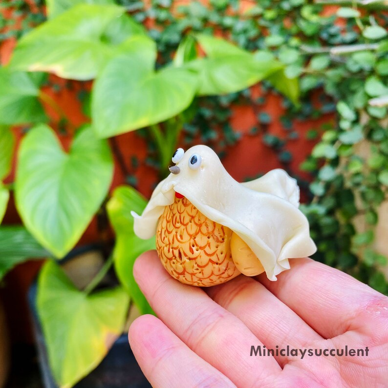 May include: A small, white ghost-shaped figurine made of clay, with googly eyes, is perched on a hand. The ghost is draped over a small, round, brown object with a textured surface.