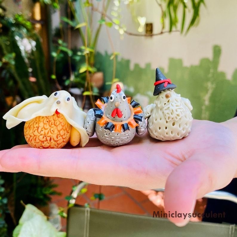 May include: Three handmade polymer clay figurines: a ghost, a chicken, and a witch. The ghost is white with a black face and is wearing a white hood. The chicken is gray with orange and black accents and is wearing a red and black collar. The witch is white with a black hat and a black beard. The figurines are sitting on a hand.