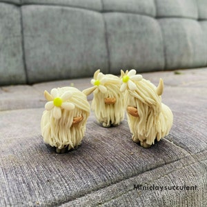 May include: Three white fluffy Highland Cow figurines with white daisy flowers on their heads. The figurines are made of polymer clay and are standing on a gray textured surface.