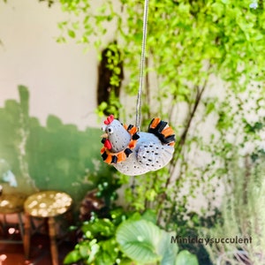 May include: A gray clay chicken ornament with orange and black accents hanging from a string. The chicken has a red comb and wattle.