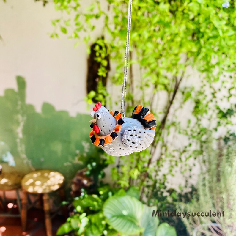May include: A gray clay chicken ornament with orange and black accents hanging from a string. The chicken has a red comb and wattle.