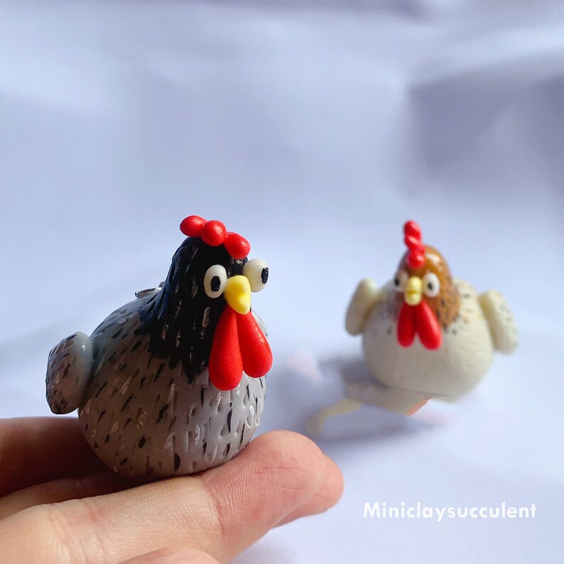 Peut inclure: Deux figurines de poulets en p&acirc;te polym&egrave;re faites &agrave; la main. L'une est grise avec des accents noirs et une cr&ecirc;te rouge, l'autre est blanche avec des accents bruns et une cr&ecirc;te rouge. Les deux ont des yeux jaunes.