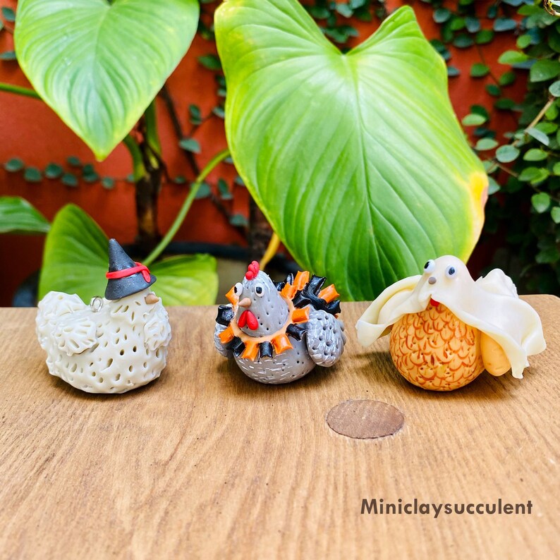 May include: Three handmade ceramic chickens decorated for Halloween. One chicken is wearing a witch's hat, another is wearing a black and orange costume, and the third is wearing a white ghost costume.