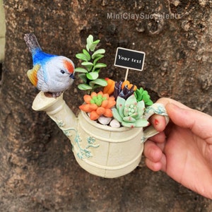 Może przedstawiać: Miniaturowy ogródek sukulentów z gliny w białym konewce z niebieskim ptakiem siedzącym na dzióbku. Ogródek zawiera różne sukulenty w odcieniach zieleni, pomarańczy i czerwieni. Mały czarny znak z tekstem "Your text" jest umieszczony w ogrodzie.