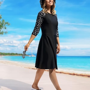 May include: A black sun dress with three-quarter sleeves featuring a white pattern. The dress is paired with a black sun hat and sunglasses. The background shows a beach with turquoise water and a blue sky.