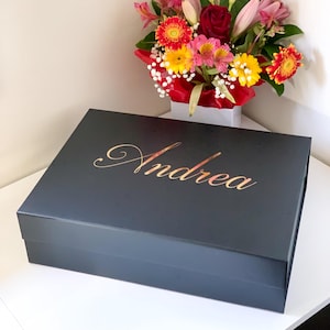 May include: A black gift box with the name "Andrea" in gold script on the lid. A colourful bouquet of flowers sits in the background. The box is on a white surface.