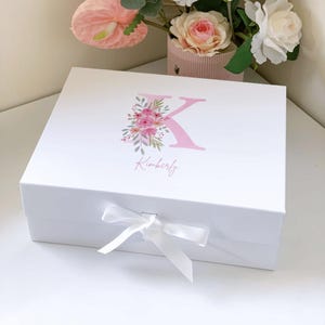 May include: A white gift box tied with a white ribbon. The lid is decorated with a pink floral arrangement around the letter 'K' and the name 'Kimberly' in script. The box is on a white surface.