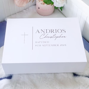 May include: A white rectangular gift box with a cross and text that reads "ANDRIOS Christopher BAPTISED 8th SEPTEMBER 2024". The box is closed and sits on a white fluffy surface. Flowers are visible in the background.