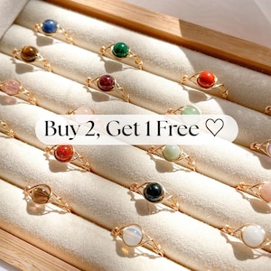 May include: A collection of gold wire rings, each featuring a different colored gemstone. The rings are arranged in a display case with the text "Buy 2, Get 1 Free" overlaid on the image.