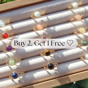May include: A collection of gold-toned wire-wrapped rings, each featuring a different colored gemstone. The gemstones include red, purple, green, blue, and other colors. The rings are displayed in a wooden box with a light-colored fabric lining. Text on the image reads "Buy 2, Get 1 Free".