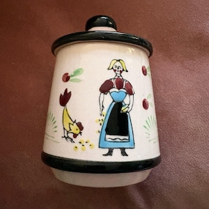 May include: A ceramic jar with a black lid and base, featuring a hand-painted design. The design includes a woman in a blue dress and a yellow rooster. Red and green accents complete the folk art style.