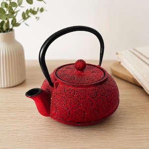 May include: A round, red cast iron teapot featuring a textured floral pattern across its surface. The teapot includes a black arched handle, a matching red lid with a small knob, and a short, curved spout extending from the side of the vessel.
