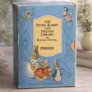 Peut inclure: Un coffret bleu vintage intitulé "The Peter Rabbit and Friends Library" par Beatrix Potter. La boîte présente des illustrations de Pierre Lapin et d'autres personnages. Le texte "The Original and Authorized Editions" est imprimé en bas.