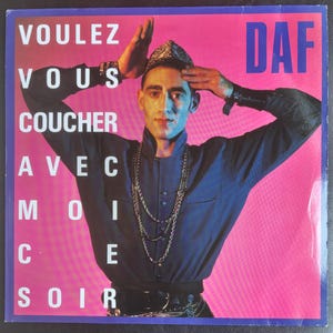 May include: A vinyl record cover featuring a person wearing a dark blue button-down shirt, a patterned headpiece, and layered silver chain necklaces. The background is a vibrant pink gradient. Text reads VOULEZ VOUS COUCHER AVEC MOI CE SOIR and DAF.