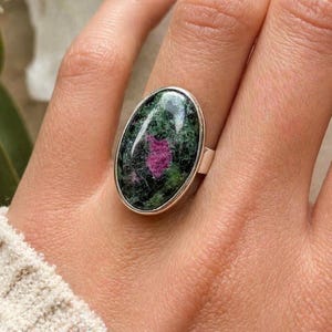 Peut inclure: Une bague en argent ornée d'une grande pierre cabochon de forme ovale à la texture marbrée. La pierre présente une base vert forêt profond avec des inclusions irrégulières et marquées de couleur magenta vif et de noir sur toute la surface.