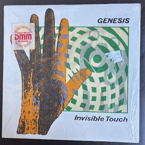 May include: Genesis "Invisible Touch" vinyl record album. The cover art displays a hand illustration in orange, purple, and black, set against a green and white backdrop. The band name and album title are printed on the cover.
