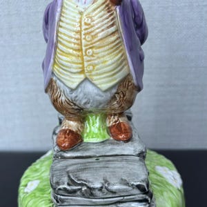 May include: A ceramic mouse figurine wearing a purple coat, yellow waistcoat, and a pink and white checked bow tie, holding a pipe. The mouse stands on a stack of books on a grassy base with white flowers.