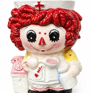 May include: A ceramic nurse figurine with a red cross on her white cap and bright red curly hair. She wears a white uniform with a red heart and a pink telephone is next to her. The figurine is painted with bright colors.