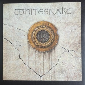May include: Album cover for Whitesnake. The band's name is carved at the top. A gold-coloured emblem with the initials "WS" is in the centre. The background has a cracked, stone-like texture. The album cover is a vintage design.