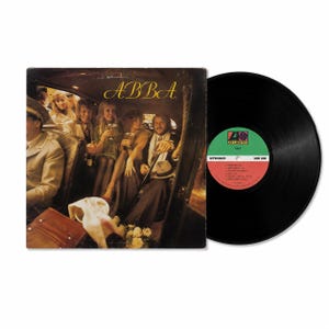 May include: A vinyl record and its square cardboard sleeve featuring the ABBA album cover. The cover shows four people sitting inside a car, with the band name ABBA in yellow text. The black vinyl record has a green and red Atlantic label.