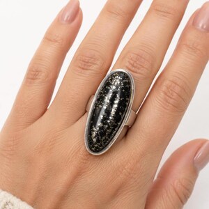 May include: A ring featuring a large, elongated oval-shaped stone with a dark black base and metallic gold-colored flecks throughout. The stone is set in a smooth, polished silver-toned metal band that encircles the finger with a simple design.