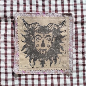 Peut inclure: Un écusson carré en tissu présentant une illustration imprimée en brun foncé d'une créature à cornes avec des cheveux fluides et des marques faciales. L'écusson a des bords dentelés et une bordure en tissu floral violet clair sur un fond vichy marron.