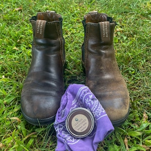 May include: A pair of brown leather boots with the brand name "Tasman Australia" on the side. A purple bandana and a tin of leather conditioner are placed in front of the boots on a bed of green grass.