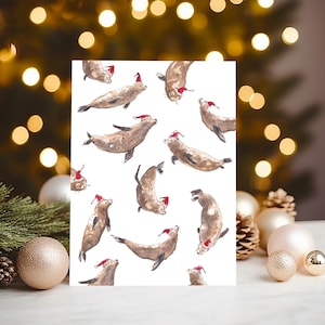 Op de afbeelding: Een witte kaart met een herhaald patroon van bruine zeehonden, elk met een rode kerstmuts. De zeehonden worden in verschillende posities afgebeeld. De achtergrond is vervaagd met kerstverlichting.