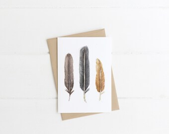 May include: A greeting card featuring three watercolor feathers in shades of brown, gray, and gold. The card is on a white background with a kraft paper envelope. The feathers are detailed and elegant.