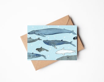 May include: A greeting card featuring an illustration of various whales in shades of blue and gray against a light blue background. The card is paired with a brown envelope, suitable for sending a message or as a decorative piece.