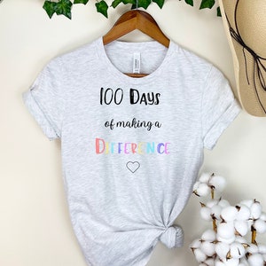 Może przedstawiać: Szary T-shirt z napisem "100 Days of making a DIFFERENCE" w kolorowych literach i małym sercem poniżej.