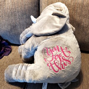 May include: A plush grey elephant toy with large ears, a trunk, and white tusks. The side of the elephant features the embroidered text "You got this" in a bright pink cursive font. The stuffed animal has a soft, fuzzy texture throughout its body.