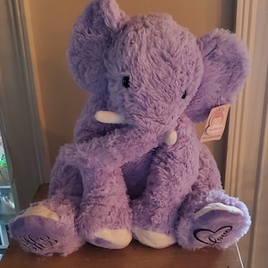 May include: A plush purple elephant toy with soft, shaggy fur, large ears, and white tusks. The left foot features an embroidered heart with the word "love" inside, while the right foot has an embroidered monogram. A tag labelled "MaoGeLan" is attached.