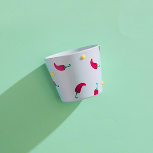 May include: A white ceramic plant pot featuring a pattern of red chilli peppers and yellow dots. The pot has a slanted design and is set against a light green backdrop, creating a shadow.