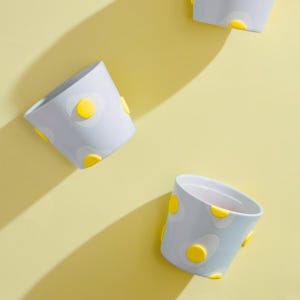 May include: Three light blue ceramic planters with a fried egg design. Each planter has a white egg shape with a yellow yolk. The planters are on a yellow background, with shadows.