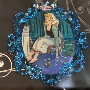 May include: A blue metal frame with ornate details surrounds a depiction of Cinderella in a blue dress, sitting on a chair and looking down at a glass slipper. The background is a teal green color.