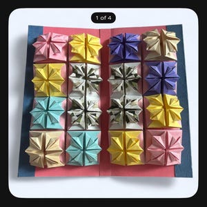 May include: A rectangular pink cardstock display featuring sixteen folded paper origami star shapes arranged in a four-by-four grid. The stars are in various colors including yellow, pink, light blue, purple, and patterned white and black.