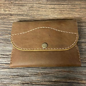 May include: A brown leather wallet featuring a curved flap closure secured with a single brass-toned metal snap button. The wallet is constructed with visible light-colored stitching along the edges of the flap and the main body of the piece.
