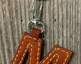 May include: A brown leather keychain with the letter "M" stitched in a contrasting light brown thread. The keychain is attached to a silver key ring.